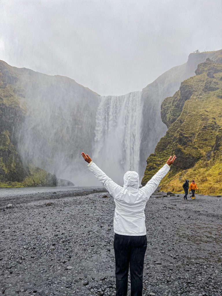 foto da Camilli usando casaco branco, de costas com braços abertos em frente a uma cachoeira no post 3 dias na Islândia 