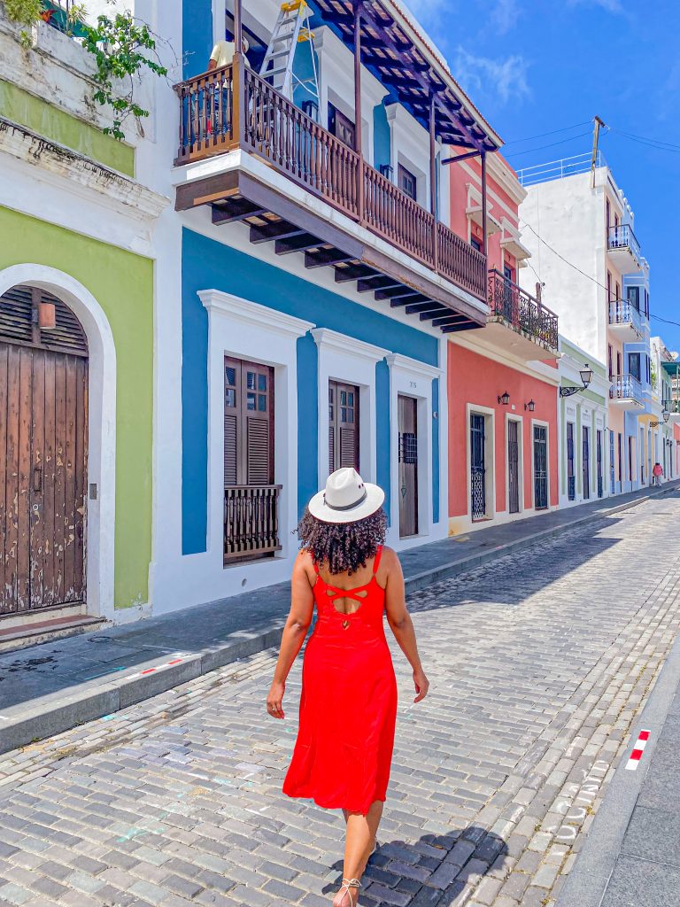 Foto da Camilli de costas, com vestido vermelho r chapéu branco no meio de uma rua de San Juan no post roteiro de 3 dias em Porto Rico
