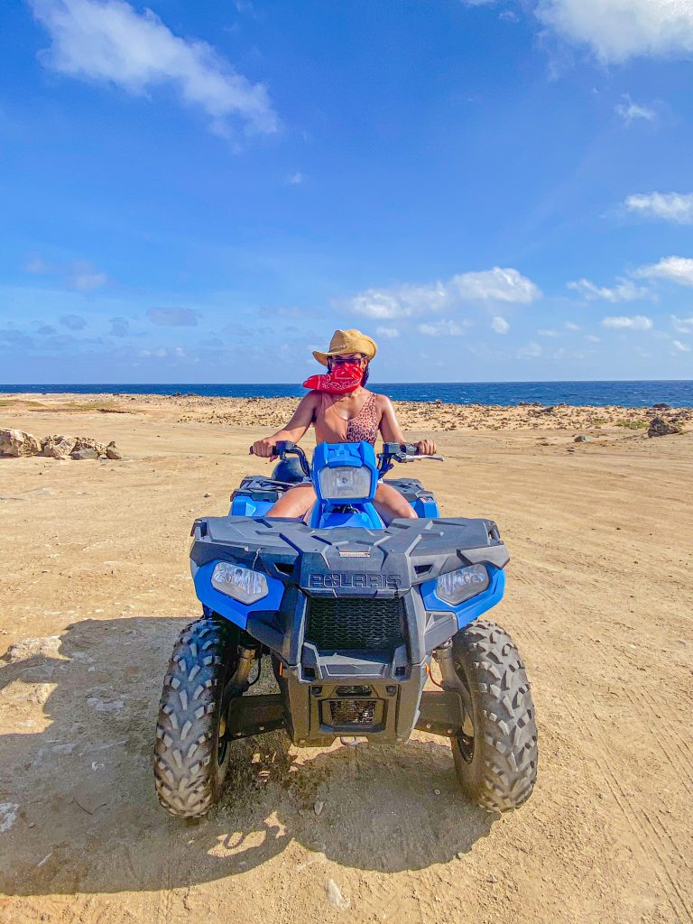 Foto da Camilli sentada em um quadriciclo azul, usando chapéu de cowboy e maio de oncinha em uma praia em Aruba