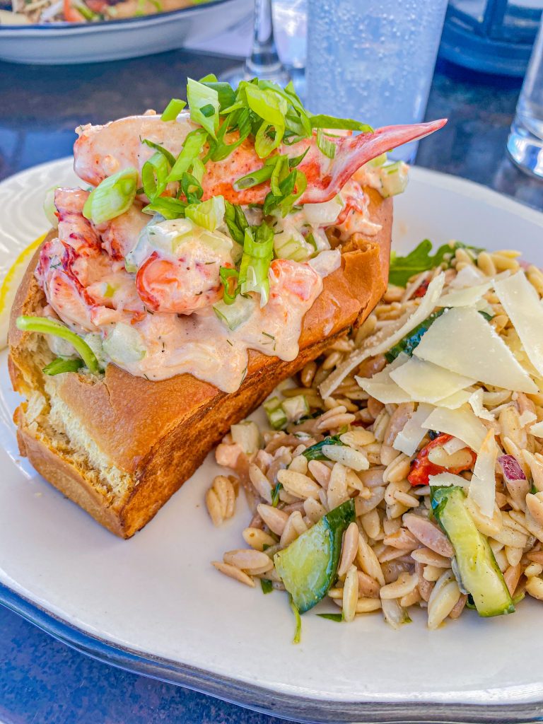 Foto de um sanduíche feito de pão de brioche e recheado com lagosta e cebolinha, e o acompanhamento é salada de macarrão com pepino, tomate, cebola e queijo parmesão 