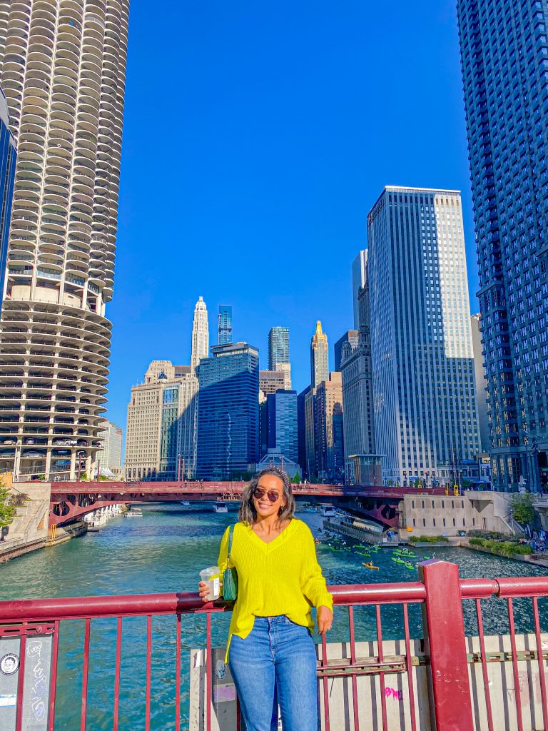 Foto da Camilli, de frente, usando um suéter verde limão, calça jeans, bolsa verde, óculos de sol e um copo do Starbucks na mão, em frente ao Rio Chicago 