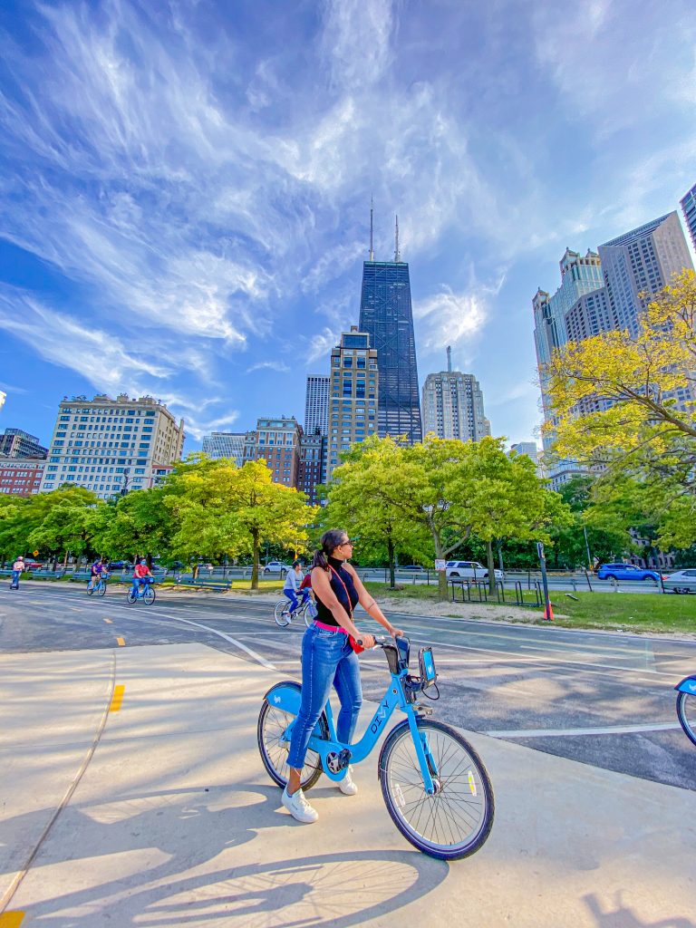 Foto da Camilli com calça jeans, tênis branco, cinto rosa e regata de gola preta, em umsa bicicleta azul em frente dos edifícios em Chicago 