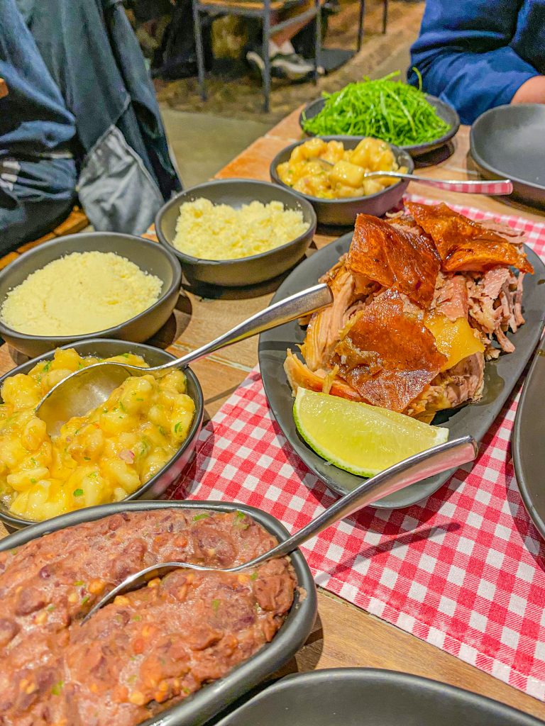 Mesa com vários pratos de comida como farofa, tutu de feijão, porco assado, banana da terra e couve no restaurante Casa do Porco