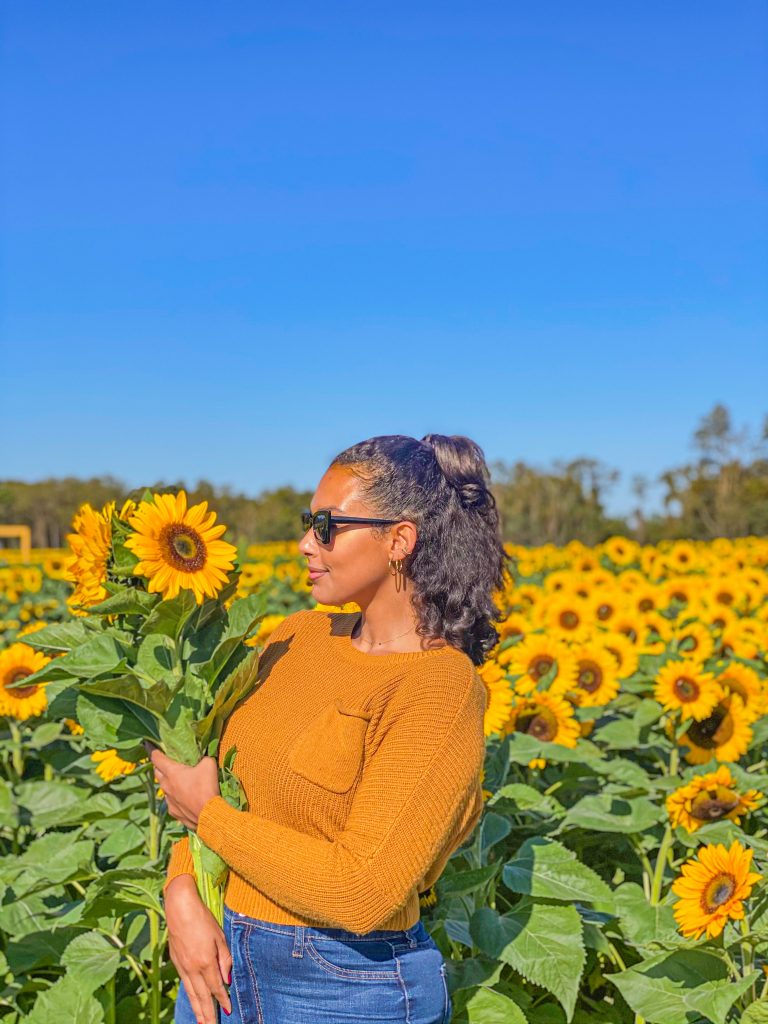 Foto da Camilli usando calça jeans e suéter amarelo, de lado segurando um girassol com vários girassóis no fundo