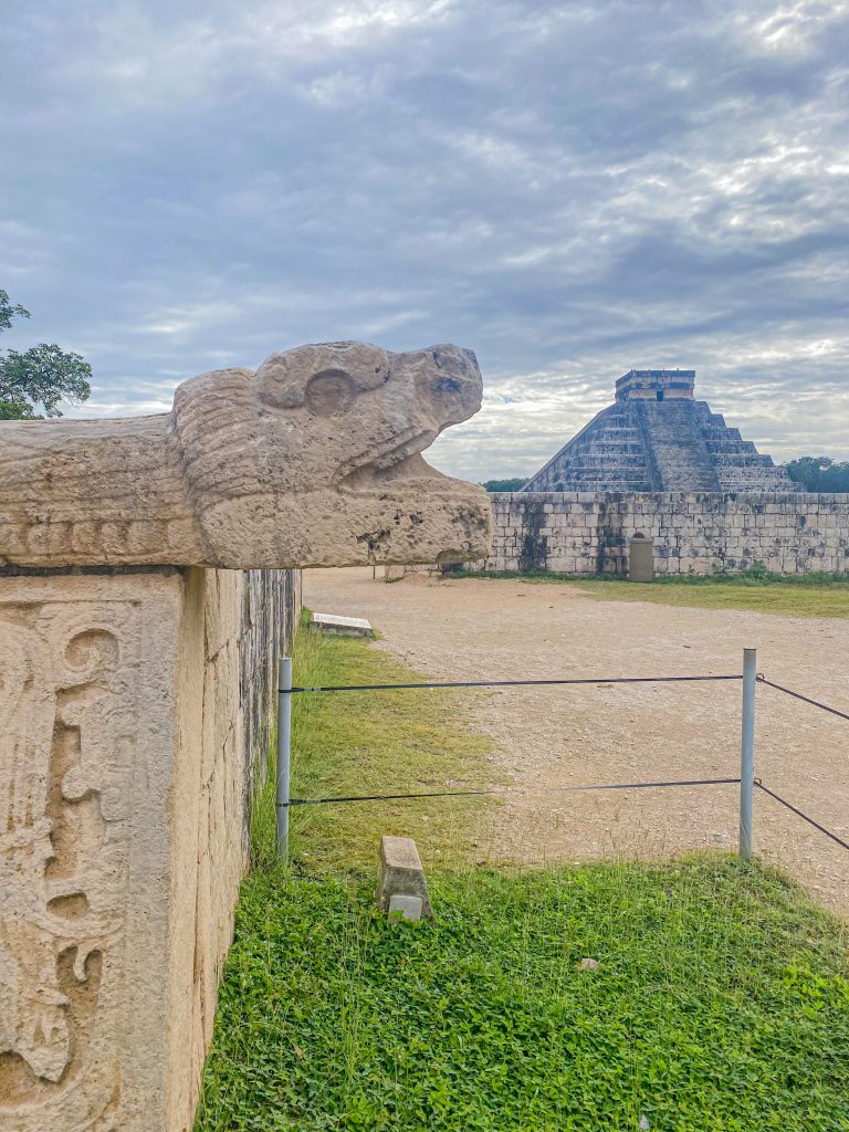 Foto se uma escultura de serpente e no fundo a pirâmide do  Chichen Itza